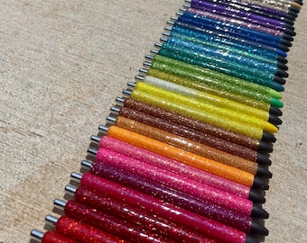 May include: A collection of glitter pens in a rainbow of colors, including red, pink, orange, yellow, green, blue, and purple. Each pen has a silver clip and a black tip. The pens are arranged diagonally, showcasing the variety of colors.