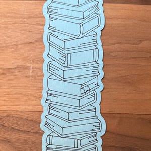 Puede incluir: Una pegatina azul claro con una pila de libros. Los libros est&aacute;n delineados en negro, con detalles como los lomos y las p&aacute;ginas visibles. La pegatina tiene un borde azul claro y est&aacute; sobre un fondo de madera.