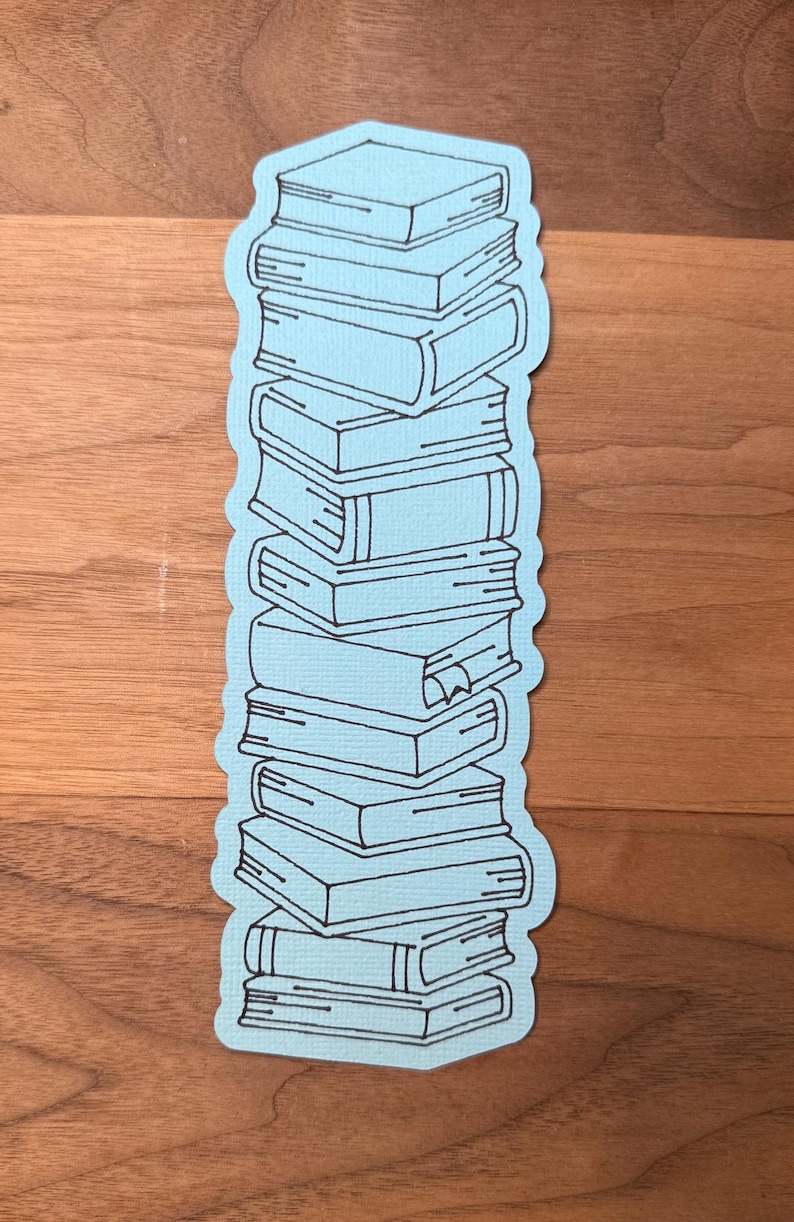 Puede incluir: Una pegatina azul claro con una pila de libros. Los libros est&aacute;n delineados en negro, con detalles como los lomos y las p&aacute;ginas visibles. La pegatina tiene un borde azul claro y est&aacute; sobre un fondo de madera.