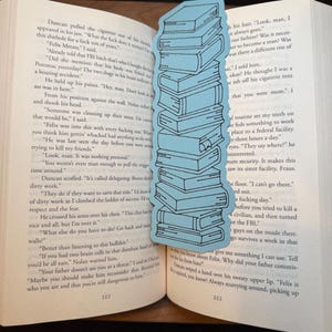 Puede incluir: Un marcap&aacute;ginas azul claro con forma de pila de libros. El marcap&aacute;ginas est&aacute; colocado en un libro abierto con texto en las p&aacute;ginas. El dise&ntilde;o del marcap&aacute;ginas es sencillo.