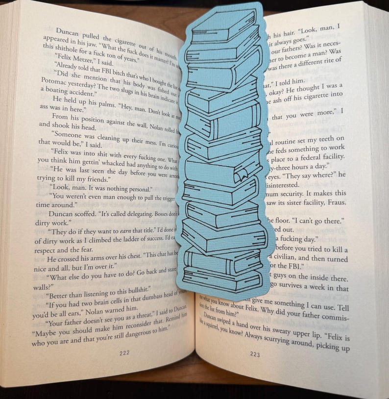 Puede incluir: Un marcap&aacute;ginas azul claro con forma de pila de libros. El marcap&aacute;ginas est&aacute; colocado en un libro abierto con texto en las p&aacute;ginas. El dise&ntilde;o del marcap&aacute;ginas es sencillo.