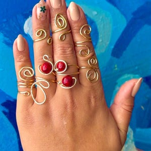 Op de afbeelding: Een hand met verschillende goudkleurige draadringen. Sommige ringen hebben een spiraalontwerp, andere hebben een rode kraal. De ringen worden op meerdere vingers gedragen en tonen diverse stijlen en vormen. De achtergrond is helderblauw.