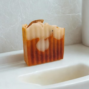 Puede incluir: Una pastilla de jabón artesanal rectangular con un diseño en capas. La capa superior es de color blanco cremoso, que pasa a un naranja claro y luego a un naranja más oscuro en la parte inferior. Una pequeña rodaja de naranja seca se encuentra en la parte superior. El jabón está sobre una superficie blanca.