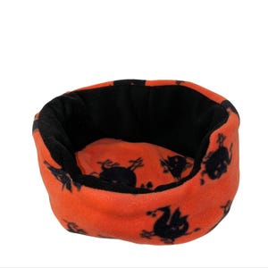 May include: A small, round pet bed with an orange exterior and a black interior. The orange fabric is patterned with black Halloween-themed silhouettes. The bed is made of a soft, plush material.