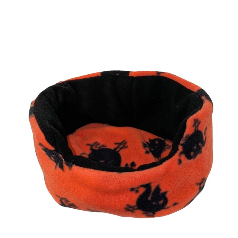 May include: A small, round pet bed with an orange exterior and a black interior. The orange fabric is patterned with black Halloween-themed silhouettes. The bed is made of a soft, plush material.