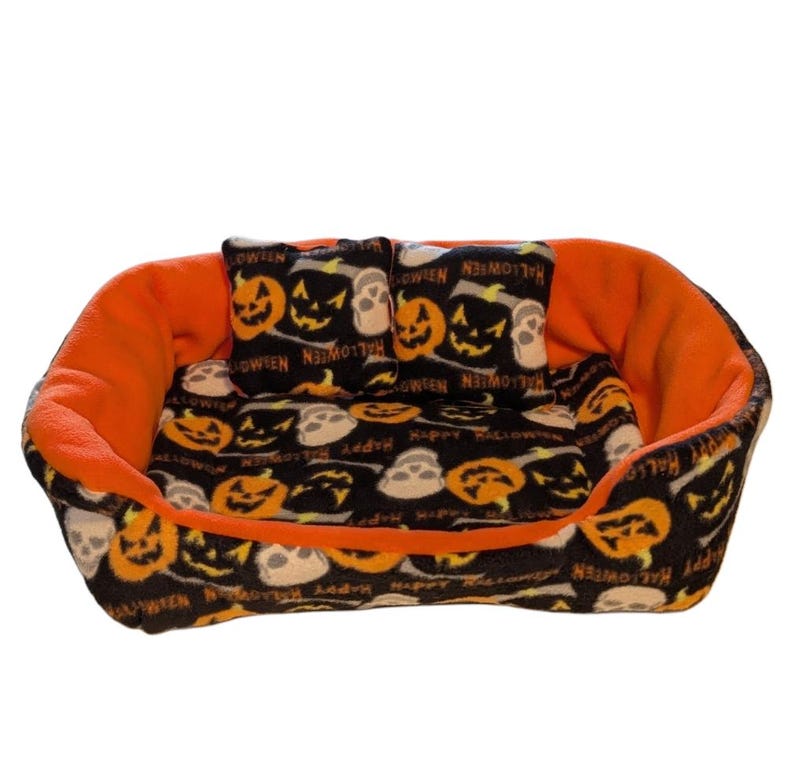 Może przedstawiać: Ł&oacute;żko dla zwierząt domowych o tematyce Halloween w kolorach czarnym i pomarańczowym. Ł&oacute;żko ma czarną tkaninę z pomarańczowymi wzorami dyni i czaszki. Wnętrze i dwie małe poduszki są czarne ze wzorami pomarańczowych dyni i czaszki. Wnętrze ł&oacute;żka jest pomarańczowe.