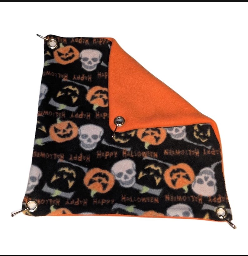 Może przedstawiać: Akcesorium dla zwierząt domowych o tematyce Halloween. Czarna tkanina przedstawia pomarańczowe dynie, białe czaszki i słowa "Happy Halloween". Druga strona jest w jednolitym pomarańczowym kolorze. Metalowe przelotki znajdują się w każdym rogu.