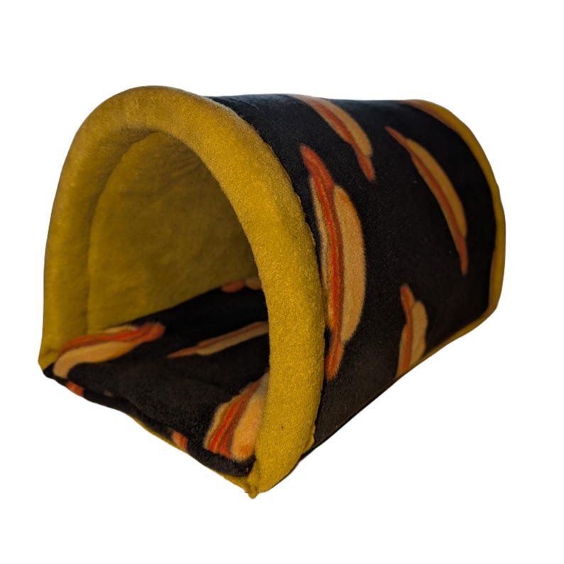 May include: A small pet tunnel bed. The interior is yellow, and the exterior is black with a pattern of orange and yellow hot dogs. The tunnel is trimmed with yellow fabric.