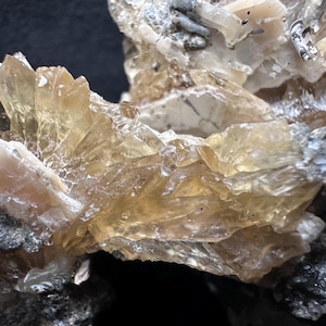May include: Close-up of a mineral specimen featuring translucent, amber-colored crystal formations. The crystals are clustered together, with a rough, textured appearance. Dark gray and white rock matrix surrounds the crystals, creating a contrast in color and texture.