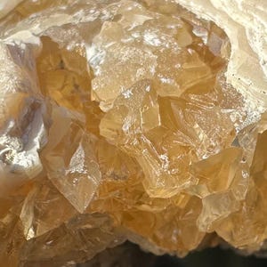 Puede incluir: Primer plano de un grupo de cristales translúcidos de color ámbar. Los cristales tienen una apariencia facetada, con bordes afilados y tamaños variables. La textura general es rugosa y cristalina, con áreas de coloración más clara, casi blanca.
