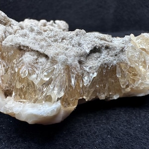 May include: A close-up of a natural mineral formation. The specimen features a crystalline structure with a light brown hue, encased in a white and grey matrix. The crystals have a translucent appearance, and the overall texture is rough and uneven.