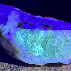 Pode incluir: Um close-up de uma formação rochosa com uma aparência em camadas. A camada superior é azul escuro, passando para um azul e verde mais claro no meio, com uma estrutura cristalina texturizada. A parte inferior é cinza claro.