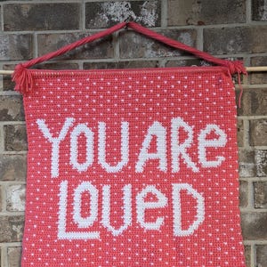 May include: A coral-colored, crocheted wall hanging with the words "YOU ARE LOVED" in white. The background is dotted with small white hearts. The banner is suspended by a pink cord and a wooden dowel.