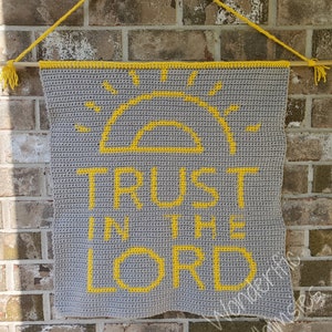 May include: A gray crochet wall hanging with yellow accents. The design features a yellow sun and the words "TRUST IN THE LORD". The banner is suspended from a wooden dowel with yellow yarn.
