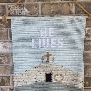 May include: A crocheted wall hanging with the words "HE LIVES" in white. The design features a church with a cross, in shades of blue, beige, and black. The banner is suspended by a wooden dowel and rope.