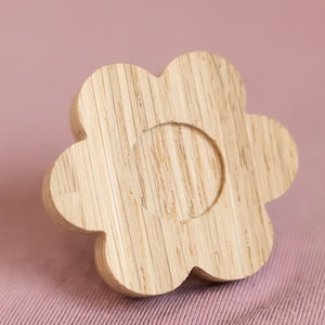 Könnte beinhalten: Ein blumenförmiger Holzknauf mit einer zentralen kreisförmigen Vertiefung. Der Knauf besteht aus hellfarbenem Holz, das die natürliche Maserung zeigt. Der Hintergrund ist ein einfarbiger rosa Stoff.