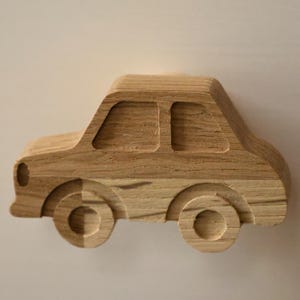 Könnte beinhalten: Ein Holzspielzeugauto mit natürlicher Holzmaserung. Das Auto hat ein klassisches Design mit zwei sichtbaren Fenstern, einer abgerundeten Front und vier geschnitzten Rädern. Das Spielzeug ist aus hellbraunem Holz gefertigt.