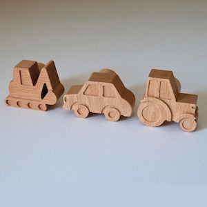 Könnte beinhalten: Drei hellbraune Wandhaken aus Holz in Fahrzeugform. Das Set besteht aus einem Bagger, einem Pkw und einem Traktor. Jedes Teil hat eine glatte Oberfläche mit natürlicher Holzmaserung und einen runden Knopf auf der Rückseite zur Befestigung.