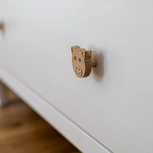 Op de afbeelding: Een lichtgekleurde houten ladeknop in de vorm van een varkenskop met gegraveerde gelaatstrekken, bevestigd aan een witte houten ladefront. De knop heeft een natuurlijke houtnerftextuur en een cilindrische basis die aan het oppervlak vastzit.