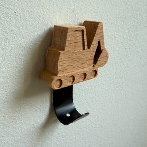 Könnte beinhalten: Ein Wandhaken mit einem hellbraunen Holzausschnitt in Form eines Baustellenbaggers mit sichtbarer Holzmaserung, befestigt an einer glatten, geschwungenen schwarzen Metallhakenbasis, die sich von der Holzfigur aus nach unten erstreckt.
