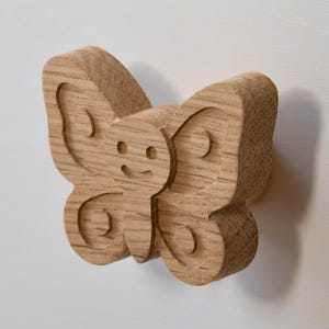 Könnte beinhalten: Eine Holzskulptur eines Schmetterlings mit einem lächelnden Gesicht und detaillierten Flügelmustern. Der Schmetterling ist aus hellem Holz gefertigt und auf einer weißen Oberfläche montiert. Die Skulptur hat einen dreidimensionalen Effekt.