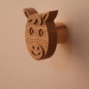 Könnte beinhalten: Ein hellbrauner Holz-Möbelknopf in Form eines Pferdekopfes. Die Vorderseite zeigt geschnitzte Details wie Ohren, eine Stirnlocke, ovale Augen und einen lächelnden Mund. Der Knopf hat einen zylindrischen Sockel, der nach außen ragt.