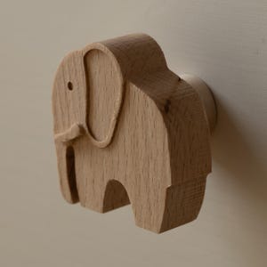 Könnte beinhalten: Ein hellbrauner Möbelknopf aus Holz in Form eines Elefanten im Profil. Der Knopf weist eine glatte, natürliche Holzmaserung mit einer kleinen kreisförmigen Aussparung für das Auge und einem geschnitzten Ohrdetail auf, montiert auf einem zylindrischen Sockel.