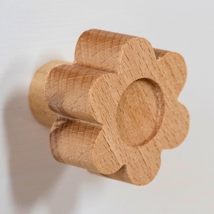 Op de afbeelding: Houten bloemvormige kastknop met een lichtbruine afwerking. De knop heeft een cirkelvormig midden en zes afgeronde bloemblaadjes. De steel is cilindrisch en bevestigd aan de achterkant van de bloem. Voor meubels.