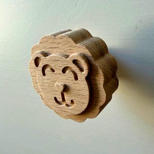 Könnte beinhalten: Ein hellbrauner Holz-Möbelknopf in Form eines Löwenkopfes. Der runde Knopf zeigt ein geschnitztes Gesicht mit zwei geschwungenen Augen, einer kleinen dreieckigen Nase und einem einfachen Mund. Die Ränder sind gewellt wie eine Löwenmähne.