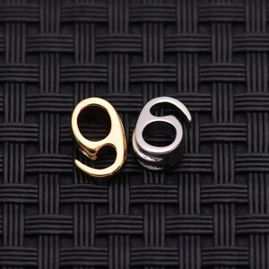 May include: Two metal charms in the shape of the number 96. One charm is gold-colored, and the other is silver-colored. The charms are displayed against a black woven background.