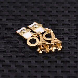 May include: Gold-toned Eiffel Tower charms and white enamel charms with a clear gem accent. The charms are connected by gold jump rings. The charms are on a black woven surface.