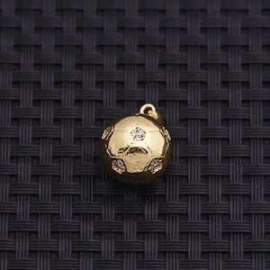 May include: A gold-colored soccer ball pendant with a small loop at the top for a chain. The ball is embellished with small, clear crystals and a star-shaped crystal on one panel. The pendant is set against a dark, textured background.