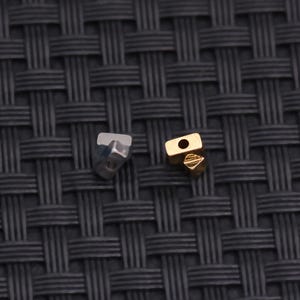 Könnte beinhalten: Nahaufnahme von zwei kleinen Metallschmuckkomponenten. Eine ist silberfarben und facettiert, die andere goldfarben und rechteckig mit einem Loch. Die Komponenten sind wahrscheinlich für die Schmuckherstellung oder -reparatur.