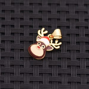 May include: A gold-colored reindeer pendant with a red and white Santa hat. The reindeer has a brown face and antlers, with a small crystal accent. The pendant is designed for jewelry.