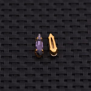 May include: Two small, teardrop-shaped jewelry pieces. One features a purple crystal set in a gold-colored metal frame. The other is a gold-colored metal finding with a curved, open design. The background is a black woven texture.