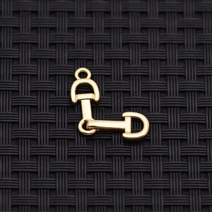 May include: A gold-colored metal charm in the shape of a horse bit, a common equestrian symbol. The charm has a ring at the top for attachment and is set against a dark, textured background.