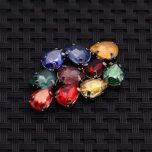 May include: A collection of colorful, teardrop-shaped rhinestones in various colors, including blue, red, yellow, and green. Each rhinestone is set in a silver-colored metal base. The rhinestones are arranged on a black woven surface.
