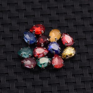 May include: A collection of colorful, teardrop-shaped rhinestones in various colors, including red, blue, green, and yellow. Each rhinestone is set in a silver-colored metal base, creating a sparkling effect. The rhinestones are arranged on a black textured surface.