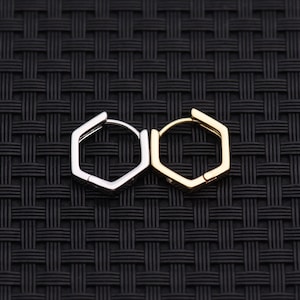May include: A pair of hexagonal hoop earrings, one silver-toned and the other gold-toned, are displayed against a dark, textured background. The earrings have a geometric design, with a hinged closure. The earrings are a modern jewelry accessory.