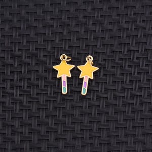 May include: Two gold-colored star-shaped charms with yellow enamel and colorful striped wands. The charms have a small loop at the top for attaching to jewelry. The background is a dark, textured surface.