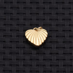 May include: A gold heart-shaped pendant with a ridged, textured surface. The pendant has a small loop at the top for attaching to a chain. The jewellery piece is set against a dark, woven background.