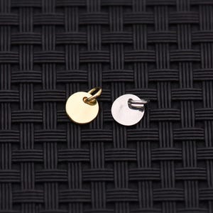 May include: Two small, round metal charms. One is gold-colored with a loop at the top, and the other is silver-colored with a small loop. The charms are displayed against a black woven background.