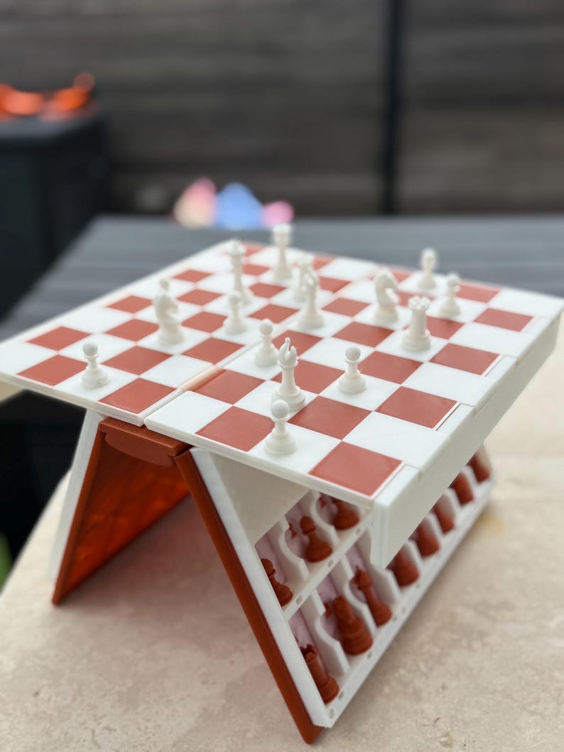 Puede incluir: Un juego de ajedrez en miniatura con un tablero a cuadros rojos y blancos y piezas blancas. El juego est&aacute; abierto, revelando un compartimento de almacenamiento para las piezas de ajedrez marrones. El juego est&aacute; sobre una superficie de color claro.