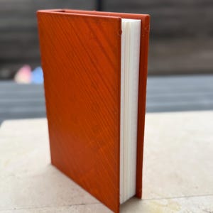 Puede incluir: Un peque&ntilde;o libro de tapa dura de color naranja con una textura de vetas de madera. Las p&aacute;ginas del libro son blancas y el lomo es visible. El libro est&aacute; de pie sobre una superficie de color claro.