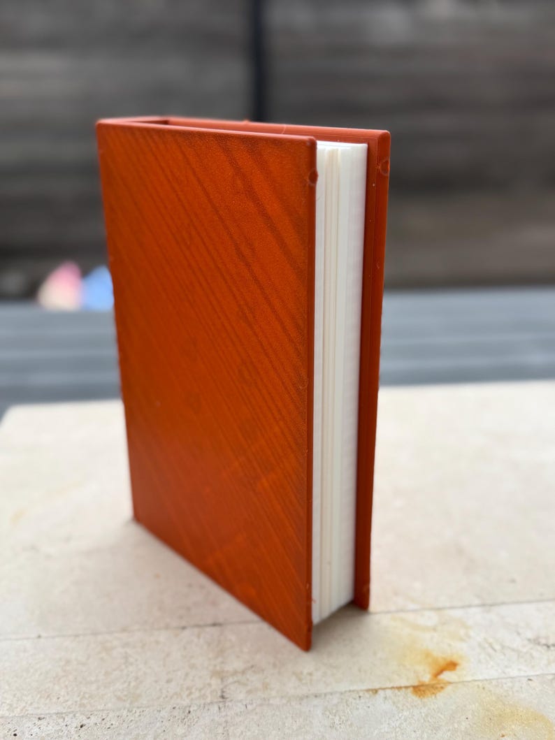 Puede incluir: Un peque&ntilde;o libro de tapa dura de color naranja con una textura de vetas de madera. Las p&aacute;ginas del libro son blancas y el lomo es visible. El libro est&aacute; de pie sobre una superficie de color claro.