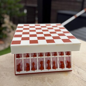 Puede incluir: Un juego de ajedrez rojo y blanco con un tablero a cuadros. Las piezas de ajedrez son rojas y se guardan en un compartimento debajo del tablero. El tablero est&aacute; abierto, revelando las piezas. El juego est&aacute; sobre una superficie clara.