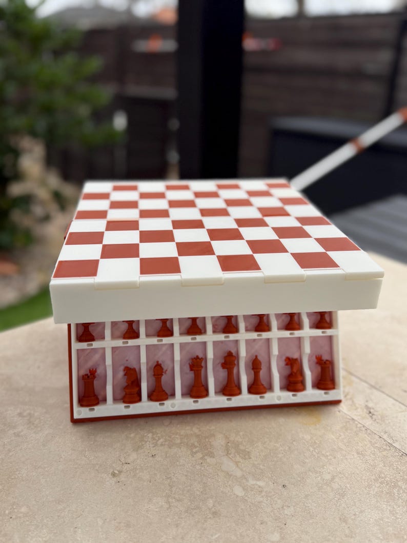 Puede incluir: Un juego de ajedrez rojo y blanco con un tablero a cuadros. Las piezas de ajedrez son rojas y se guardan en un compartimento debajo del tablero. El tablero est&aacute; abierto, revelando las piezas. El juego est&aacute; sobre una superficie clara.