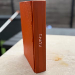 Puede incluir: Una caja de ajedrez naranja con la palabra "CHESS" impresa verticalmente en blanco. La caja rectangular est&aacute; cerrada y tiene una superficie texturizada. La caja parece estar hecha de un material duro.