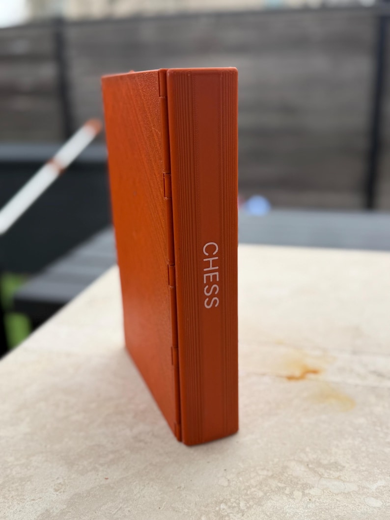Puede incluir: Una caja de ajedrez naranja con la palabra "CHESS" impresa verticalmente en blanco. La caja rectangular est&aacute; cerrada y tiene una superficie texturizada. La caja parece estar hecha de un material duro.