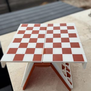Puede incluir: Un peque&ntilde;o juego de ajedrez plegable con un tablero a cuadros rojos y blancos. El tablero est&aacute; sostenido por patas blancas y naranjas. Dise&ntilde;ado para la portabilidad, es ideal para viajes o espacios reducidos. Mide aproximadamente 25 cm x 25 cm.